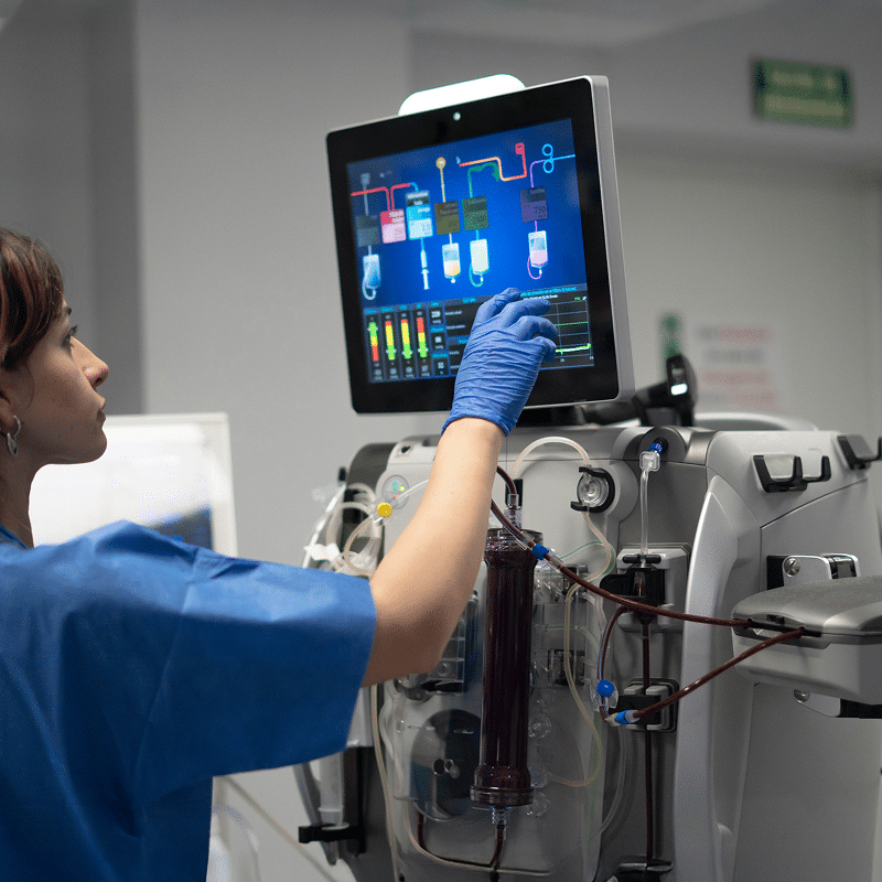Professionnel de santé en blouse bleue et gants manipule l'écran tactile d'un dialyseur. L'interface affiche des données médicales; tubes de sang visibles.